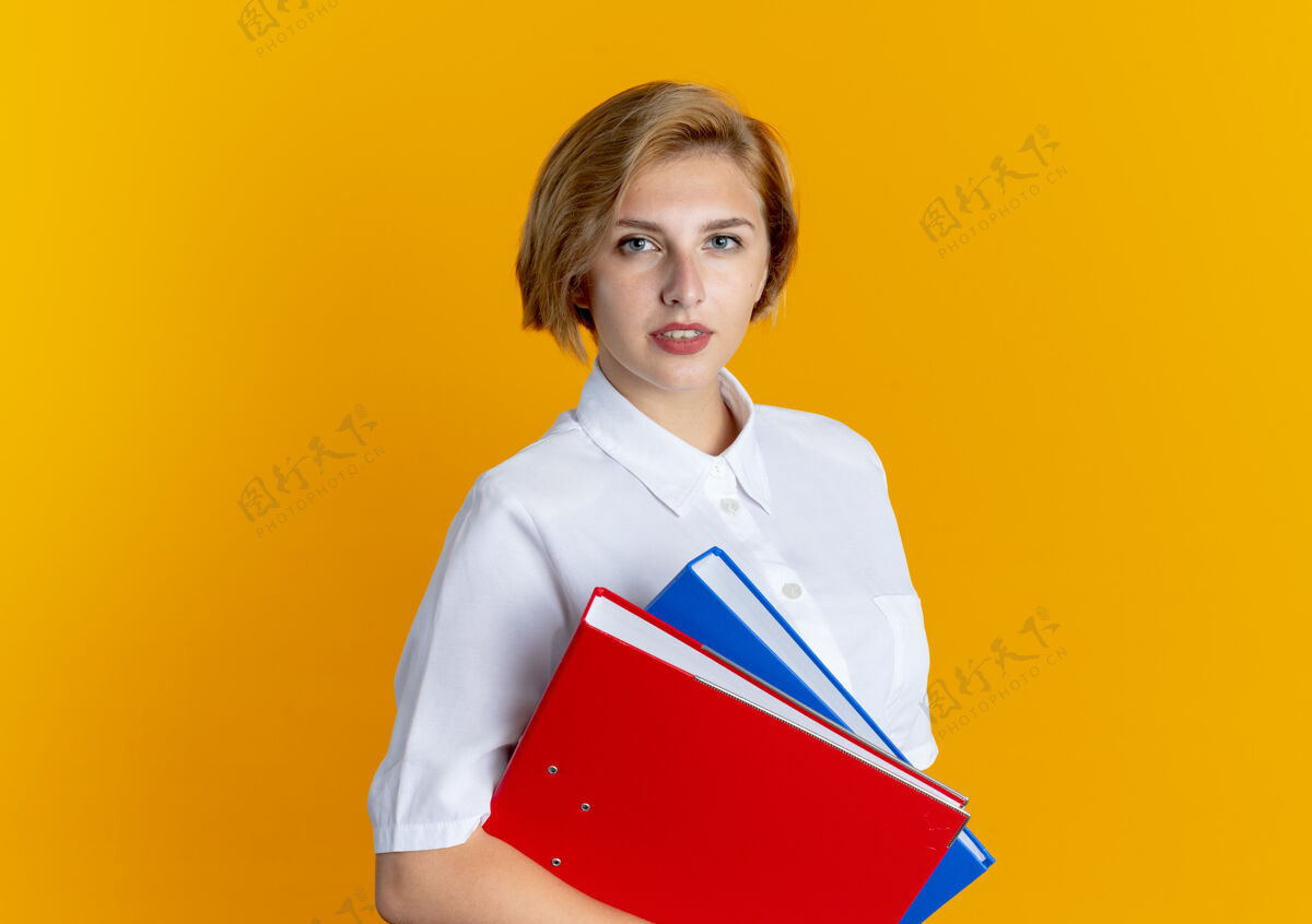 文件年轻自信的俄罗斯金发女孩拿着文件夹 看着隔离在橙色背景和复印空间的相机年轻信心相机