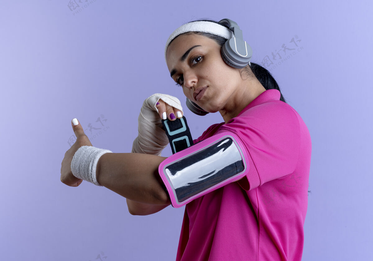 臂带年轻自信的白人运动女性戴着头带和腕带的耳机纠正手机袖带孤立的紫色背景与复制空间腕带正确耳机