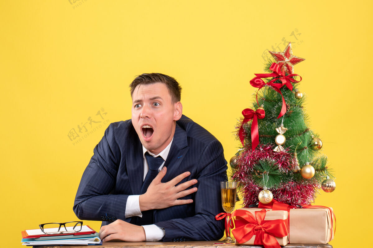前面前视图激动的年轻人抱着胸脯坐在圣诞树旁的桌子旁 呈现黄色背景胸生意人近