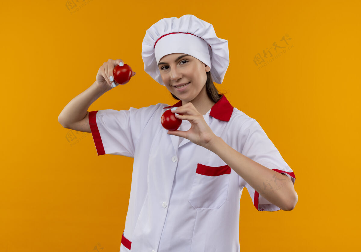 烹饪身着厨师制服 面带微笑的白人年轻厨师女孩拿着隔离在橙色背景上的西红柿 并留有复印空间制服女孩微笑