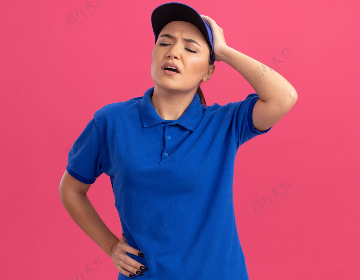 手身穿蓝色制服 戴着帽子的年轻送货员 一脸困惑 手放在头上 因为她站在粉红色的墙上犯了个错误头困惑立场