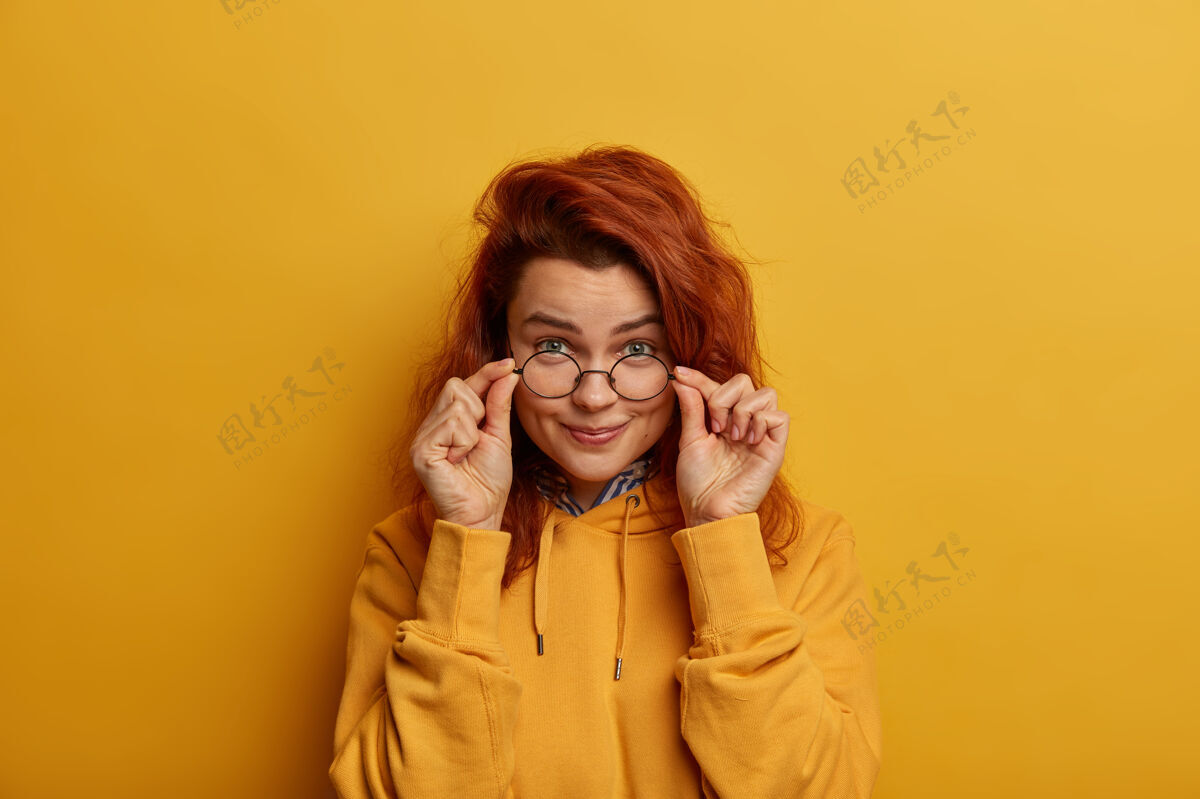 女性红发女人透过眼镜一丝不苟地看着 好奇地凝视着 手放在眼镜架上 穿着黄色的运动衫运动衫温柔白日梦