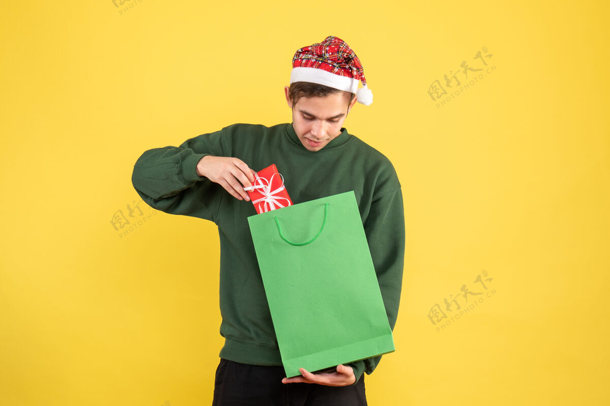 黄色正面图戴着圣诞帽的年轻人拿着绿色购物袋和礼物看着黄色背景上的礼物绿色人背景