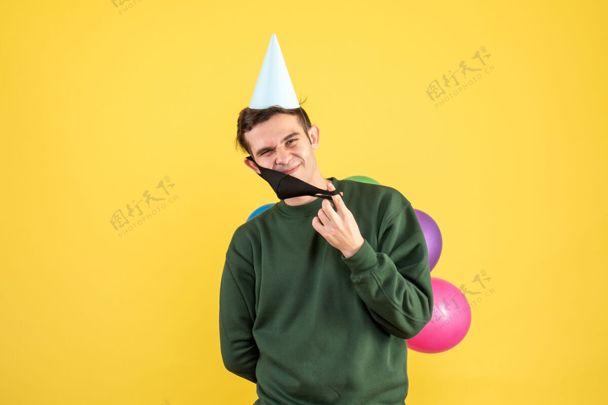 聚会正面图：戴着派对帽和彩色气球的年轻人摘下黄色背景上的面具帽子拿着背景
