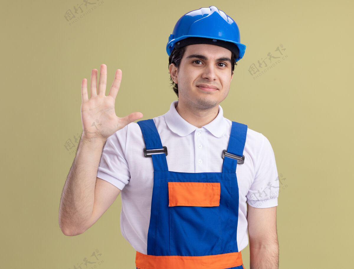 脸年轻的建筑工人穿着建筑制服 戴着安全帽 面带微笑 站在绿色的墙上挥手致意安全微笑男人