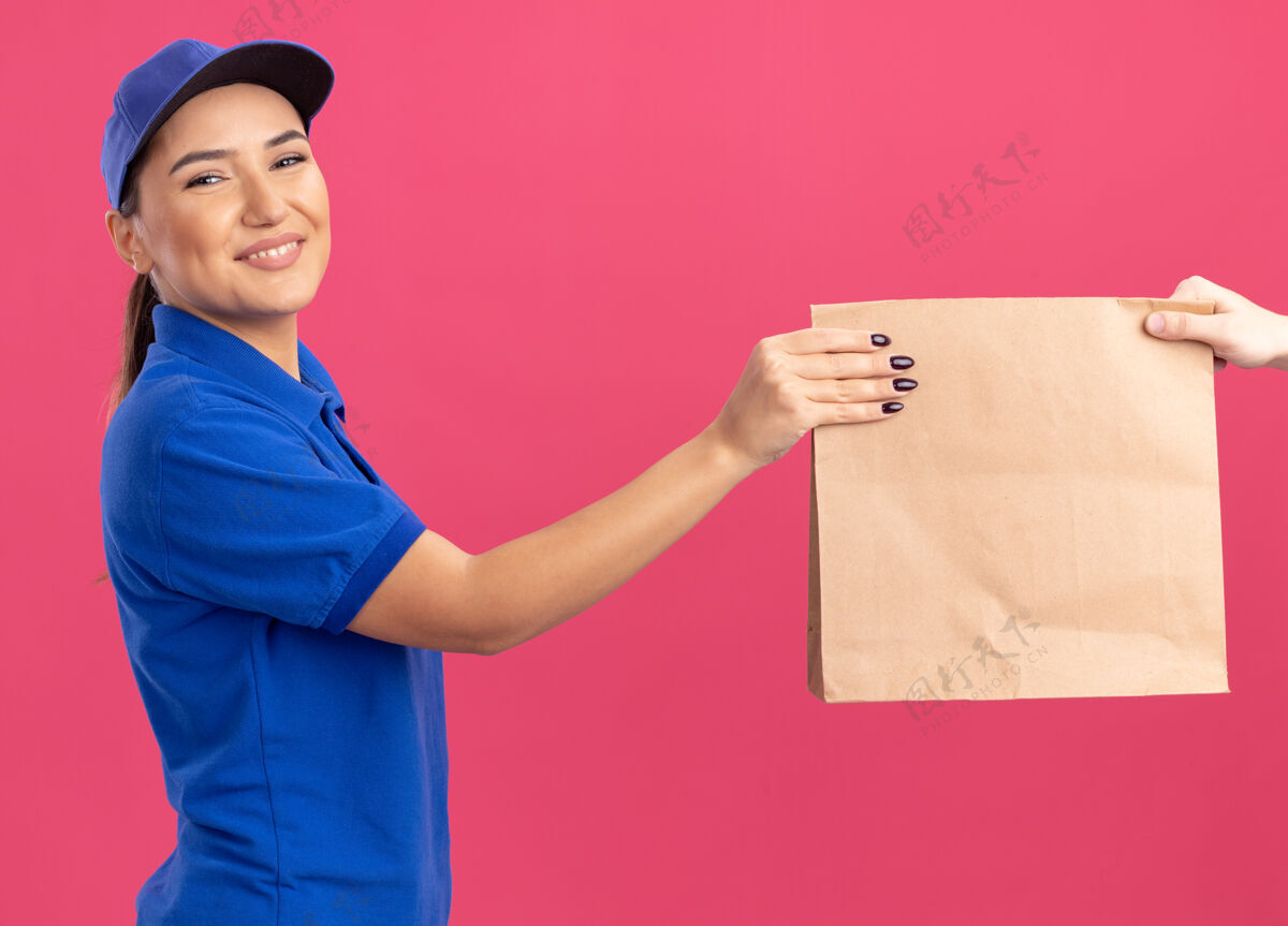 包装身着蓝色制服 头戴帽子的年轻送货员站在粉色的墙上 微笑着向顾客赠送纸质包装送货帽子顾客