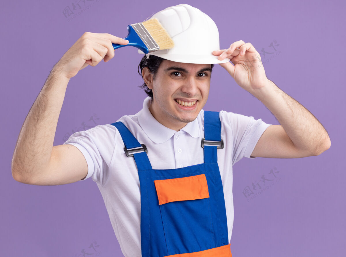 抱着快乐的年轻建筑工人穿着建筑制服 戴着安全帽 拿着油漆刷 站在紫色的墙上微笑着看着前方制服年轻人微笑