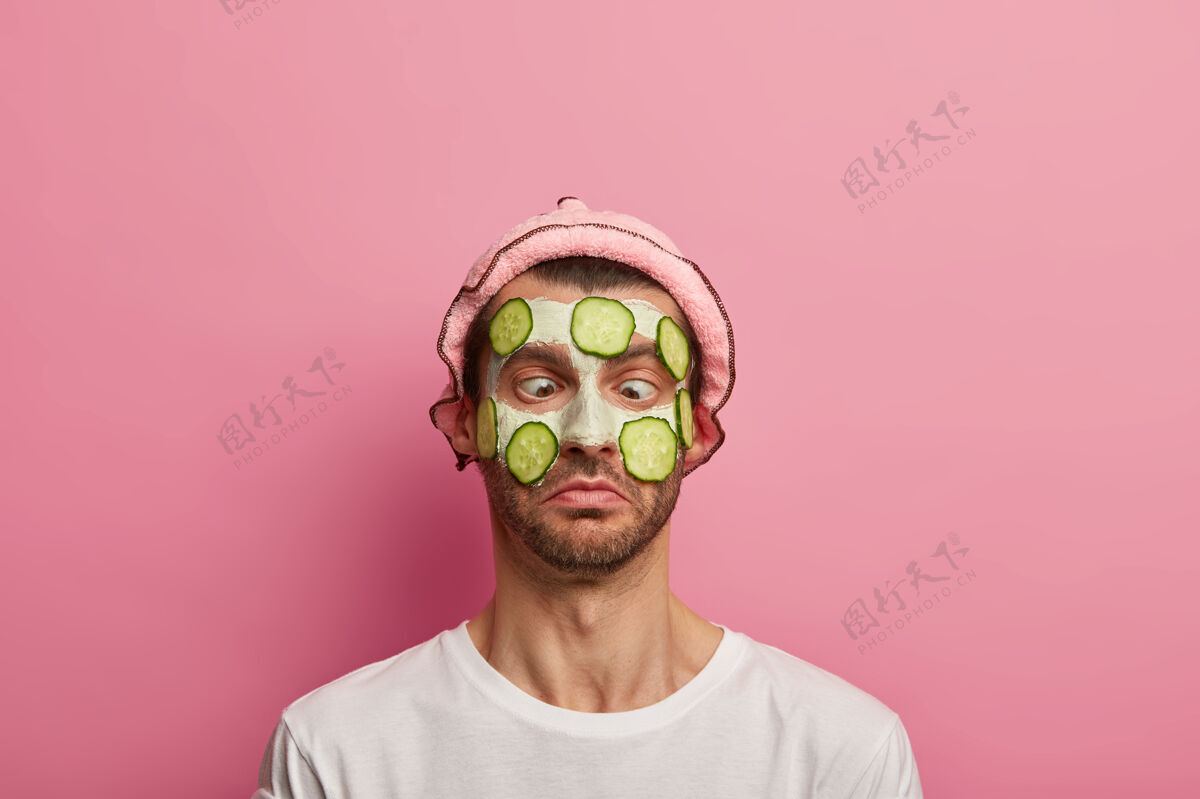 过程搞笑的男模表情滑稽 眼睛交叉 脸上戴着泥面具和黄瓜片 头戴柔软的帽子皮肤治疗T恤
