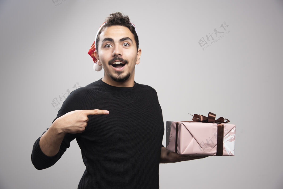 年轻人一个戴着圣诞老人帽子指着礼物的快乐男人蝴蝶结礼物圣诞老人