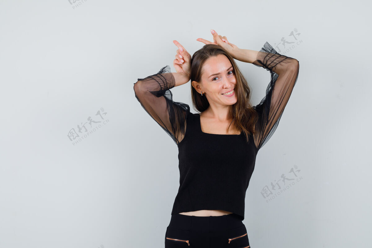 女性年轻女子举起双手 展示着兔子的姿势 穿着黑色上衣和黑色裤子 看起来很有趣 正面图手势年轻脸