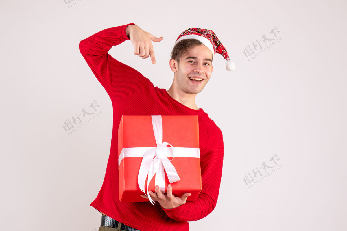 微笑前视图：戴圣诞帽的年轻人指着白色背景上的礼物可爱帽子快乐