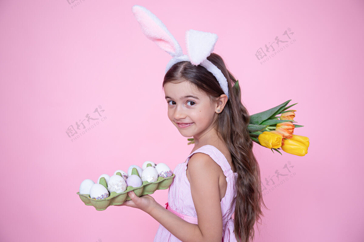 托盘有复活节兔子耳朵的小女孩手里拿着一束郁金香和一盘鸡蛋放在粉红色的墙上女孩春天兔子
