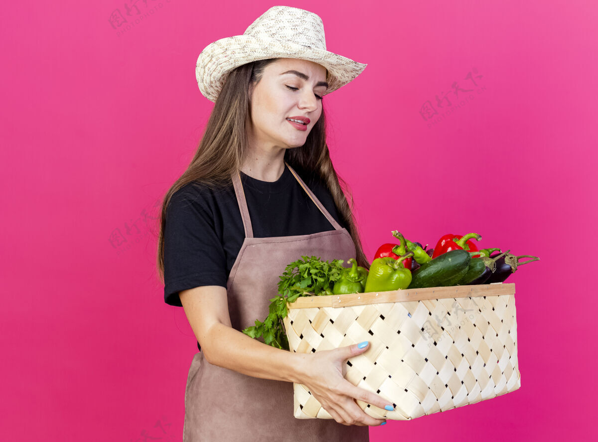 花园高兴美丽的园丁女孩穿着制服 戴着园艺帽 拿着蔬菜篮子看着孤立的粉红色背景请穿篮子拿着
