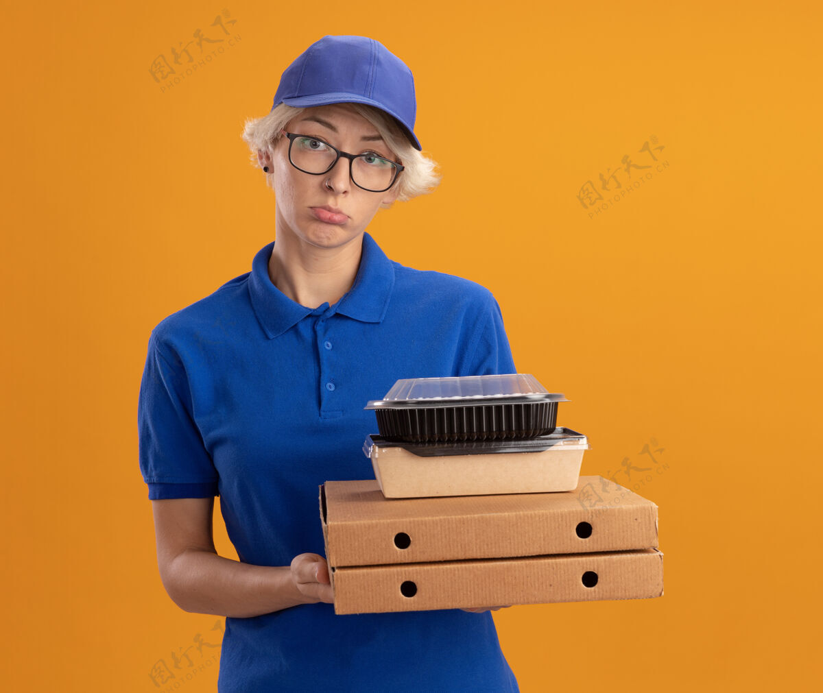 盒子身着蓝色制服 戴着眼镜 手持比萨饼盒和食品包装的年轻送货员 脸上带着悲伤的表情 越过橘色的墙壁脸表情眼镜