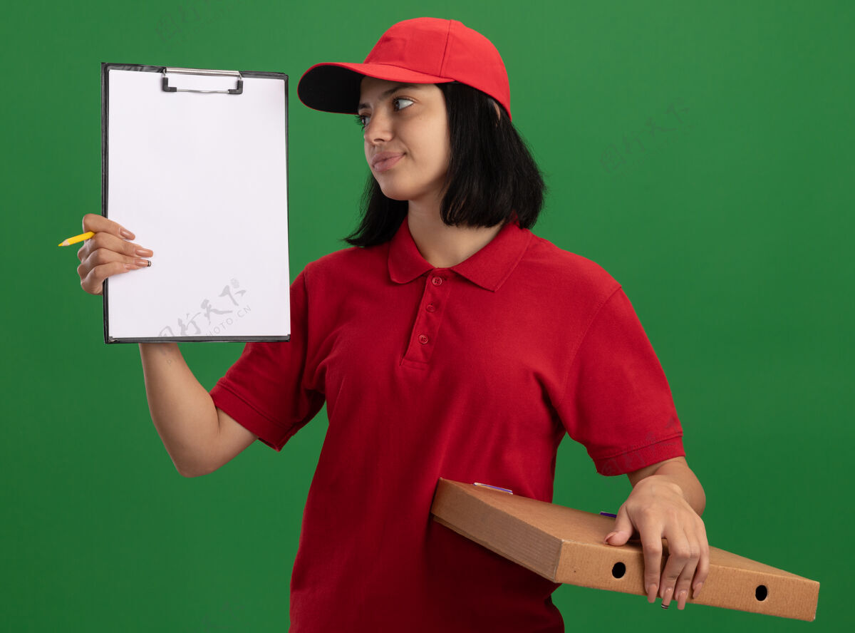 帽子身穿红色制服 头戴鸭舌帽的年轻送货女孩拿着披萨盒和铅笔的剪贴板 面带微笑地看着它站在绿色的墙上年轻送货站着