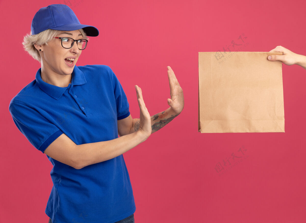 包装身穿蓝色制服 戴着帽子的年轻送货员站在粉色的墙上拒绝拿纸包裹女人制服送货