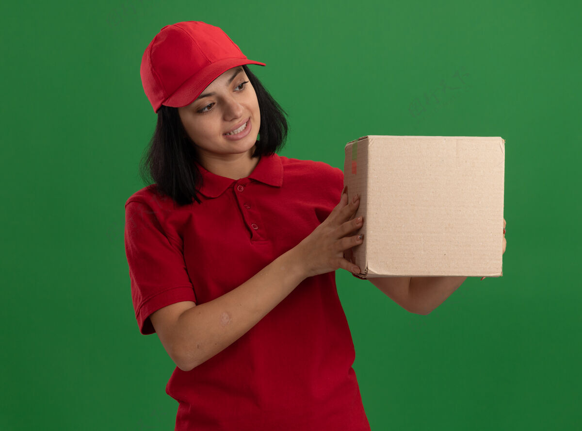 立场年轻的送货员穿着红色制服 戴着帽子 手里拿着一个纸板箱 站在绿色的墙边好奇地看着它举行帽子制服