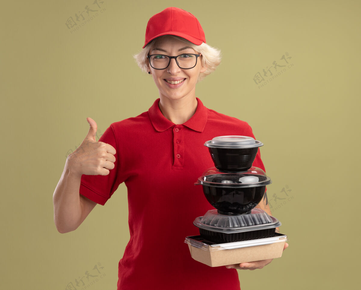 盒子快乐的年轻女送货员身穿红色制服 戴着帽子 戴着眼镜 手里拿着一叠食品包 微笑着 自信地竖起大拇指站在绿色的墙上送货自信眼镜