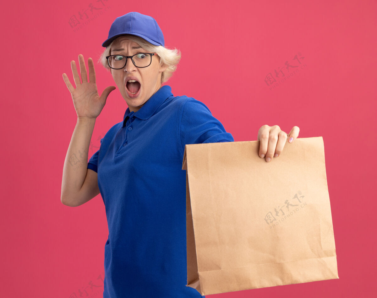交货年轻的送货员身穿蓝色制服 戴着眼镜 手里拿着纸包 惊恐地看着它 手翻在粉红色的墙上穿帽子包装