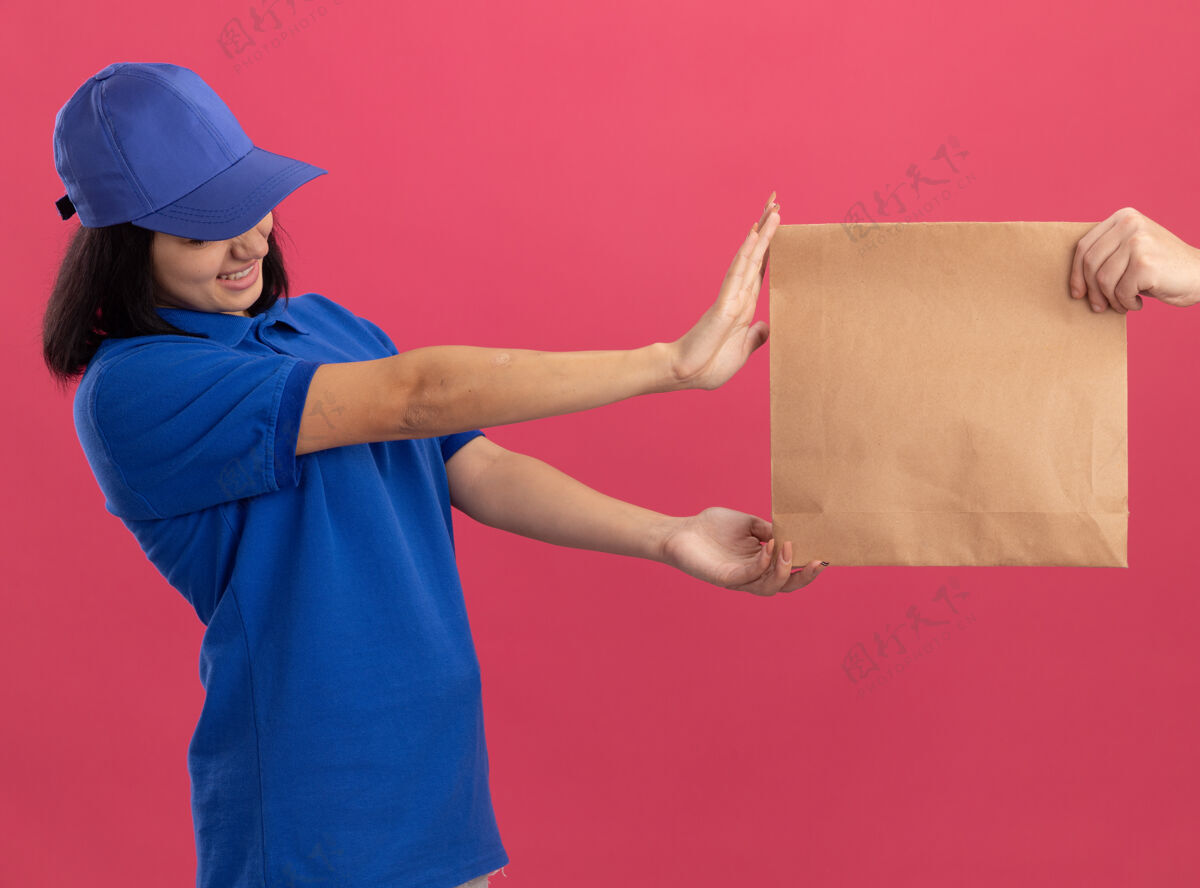 制服穿着蓝色制服 戴着帽子的年轻送货女孩站在粉红色的墙上拒绝拿纸包裹拿着女孩送货