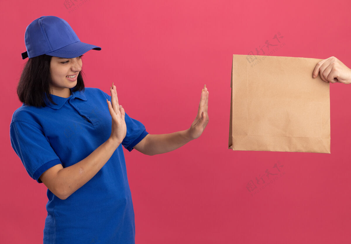 制服穿着蓝色制服 戴着帽子的年轻送货女孩站在粉红色的墙上拒绝拿纸包裹站着女孩拒绝