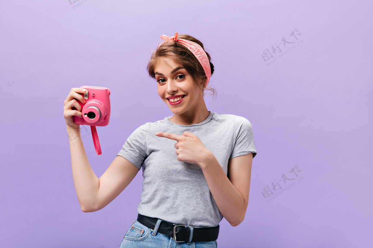 紫色穿着灰色衬衫的快乐女孩指着粉色相机漂亮可爱的女人 发型时尚 穿着灰色t恤和牛仔裙 摆出宽腰带的姿势时尚女人微笑