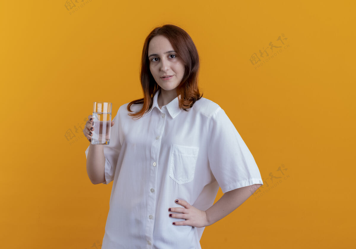 红发看着镜头 年轻的红发女孩拿着一杯水 把手放在屁股上 隔离在黄色的背景上臀部女孩相机
