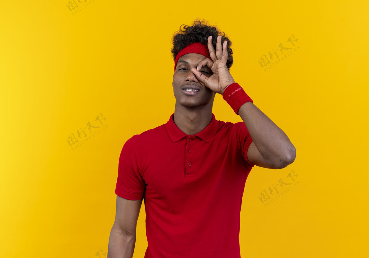 非裔美国人高兴的年轻黑人美国运动男子戴着头带和腕带显示出看手势隔离在黄色背景穿请表演