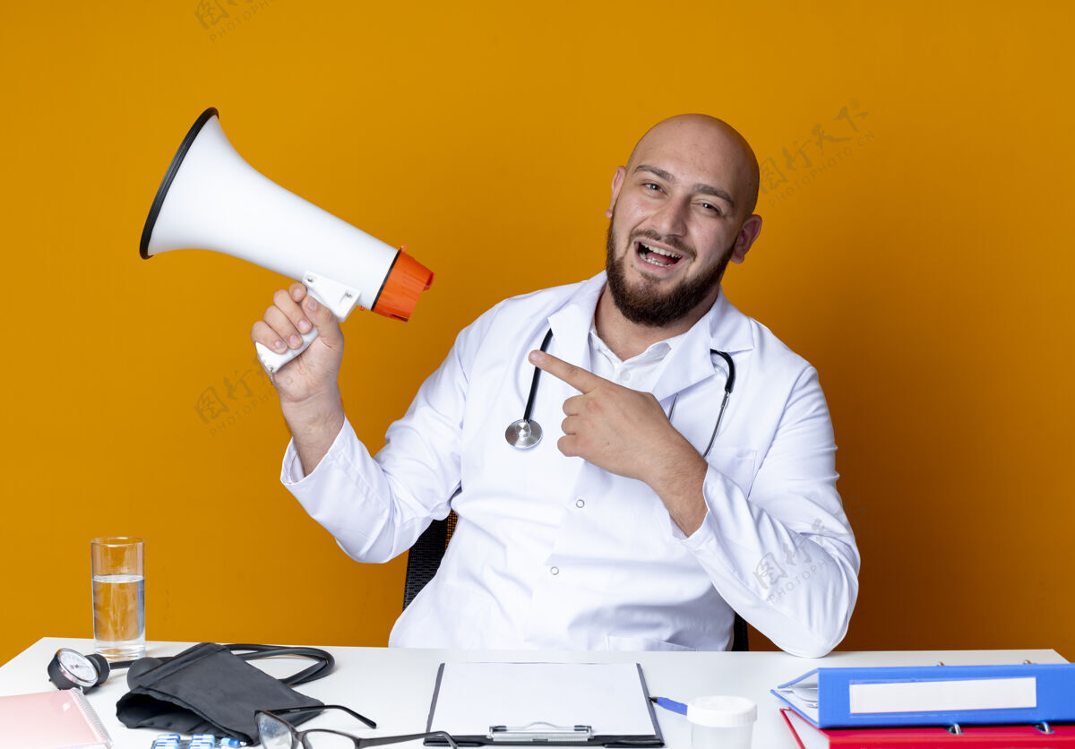 点快乐的年轻秃头男医生穿着医用长袍和听诊器坐在办公桌旁 手持医疗工具 指着橙色背景上孤立的扬声器年轻举行长袍