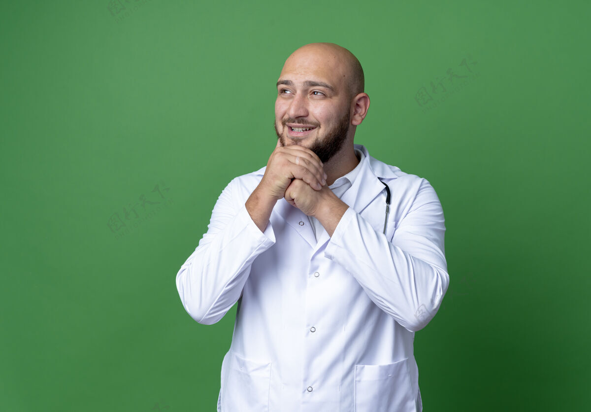 医生看着旁边高兴的年轻男医生穿着医用长袍和听诊器把双手放在下巴下隔离在绿色的背景上听诊器请穿长袍