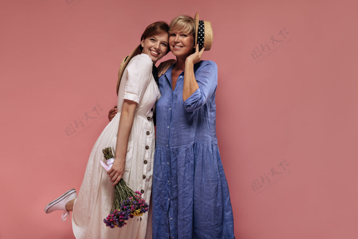 女性现代的两位女士 留着酷酷的短发 穿着时髦的连衣裙 微笑着 拥抱着 在孤立的背景上拿着花束和草帽老年女人年轻