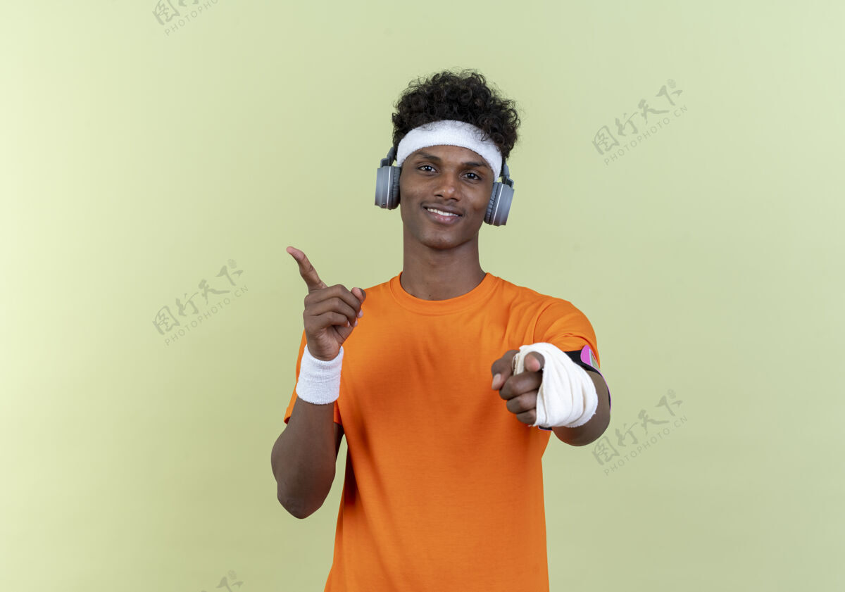 腕带高兴的年轻黑人美国运动男子戴头带和腕带和电话臂带耳机显示您的姿态和点在绿色背景与复制空间隔离侧年轻穿手臂
