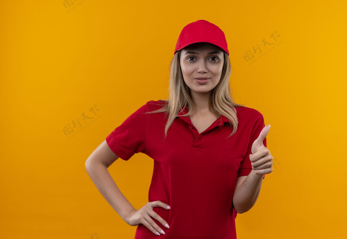 帽子穿着红色制服 戴着帽子 手放在臀部 拇指孤立地竖立在橙色背景上的年轻送货女孩很高兴请穿制服手
