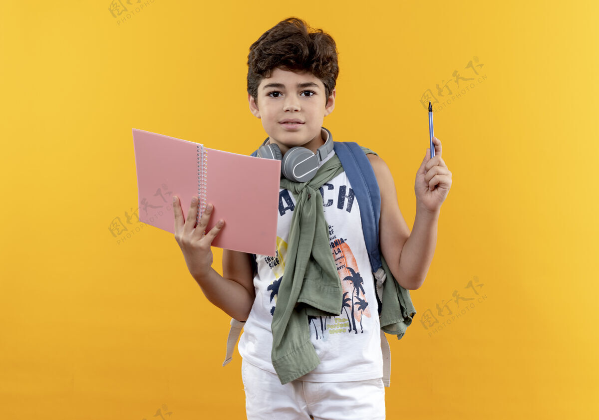 包看着摄像机 一个戴着背包和耳机的小男孩 手里拿着笔记本和笔 隔离在黄色背景上背笔抱
