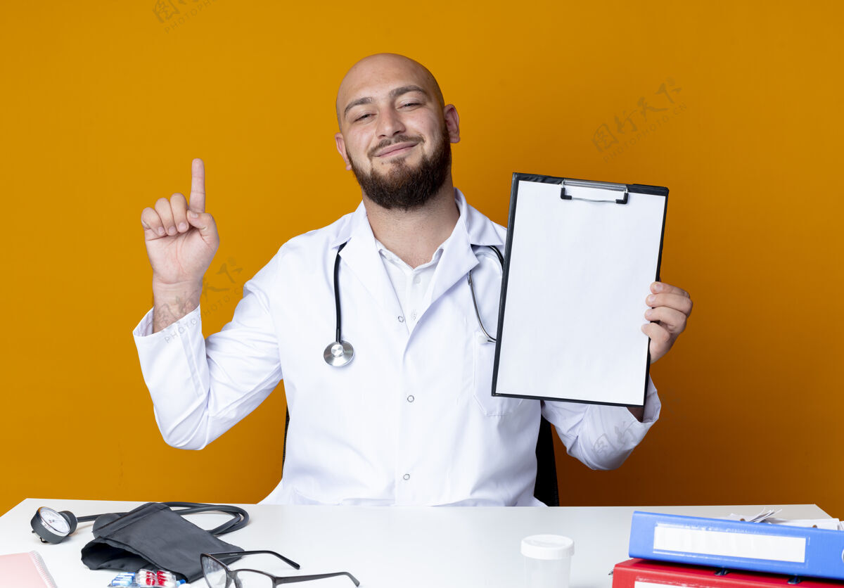 剪贴板高兴的年轻秃头男医生穿着医用长袍和听诊器坐在工作台上拿着医疗工具拿着剪贴板 指着上面孤立的橙色背景年轻秃头穿