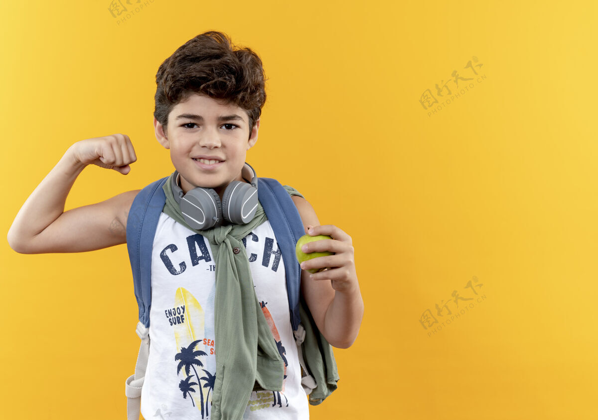 戴着高兴的小男孩戴着书包和耳机拿着苹果和显示是的手势隔离在黄色背景与复制空间拿着展示苹果