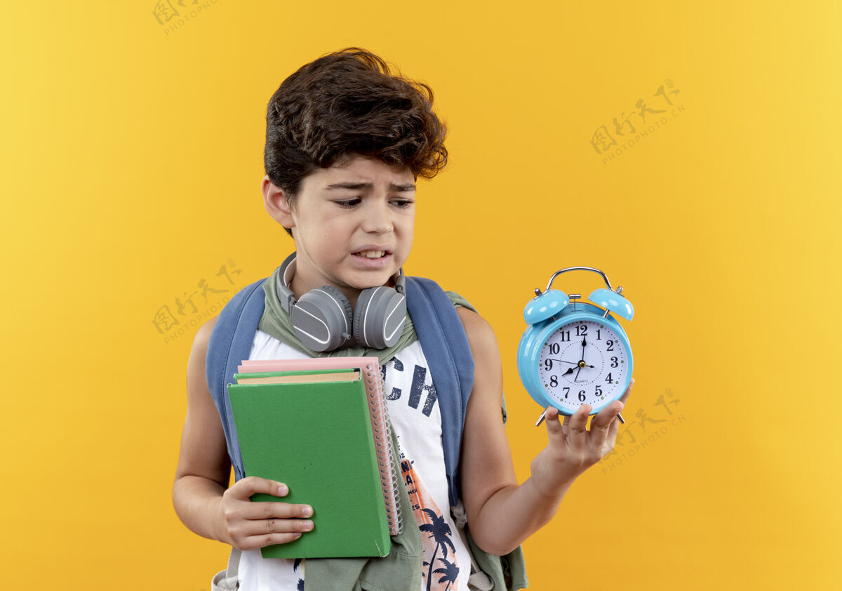 男孩关心的小男孩戴着书包和耳机拿着书 看着闹钟在他的手上孤立的黄色背景手背小
