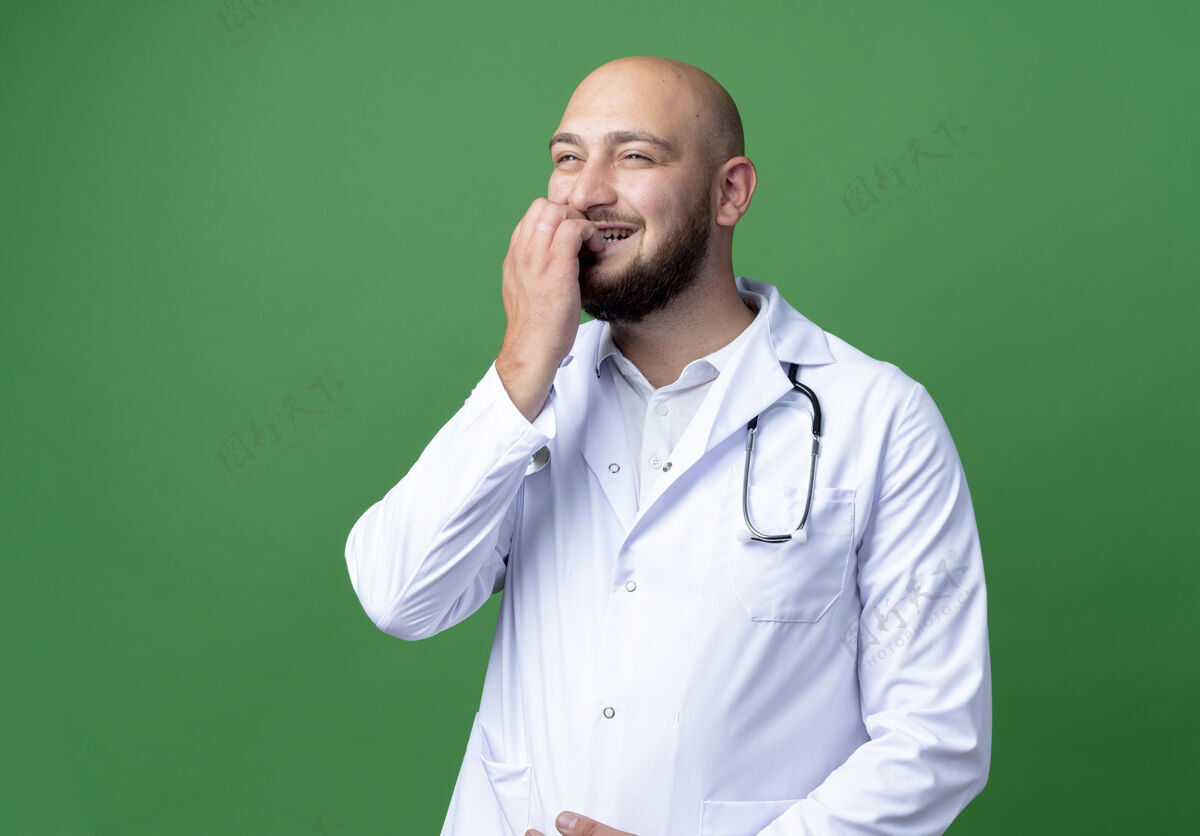 医生看着身边高兴的年轻男医生穿着医用长袍和听诊器咬指甲隔离在绿色背景上请长袍咬