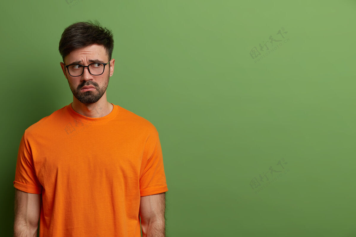 面部闷闷不乐的欧洲年轻人看起来心烦意乱 戴着休闲的橘色t恤和眼镜 心情不安 情绪低落 站在绿色的墙壁上 为你的晋升腾出空间忧郁情绪橙色