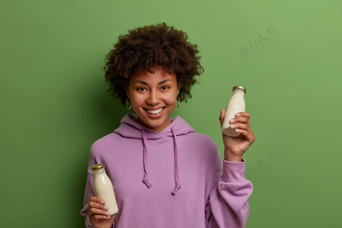 微笑快乐的素食女性 手拿玻璃瓶植物基无乳糖牛奶 有健康的营养 穿着紫色连帽衫 笑容灿烂 隔着绿色的墙壁非乳制品替代饮料室内牛奶椰子