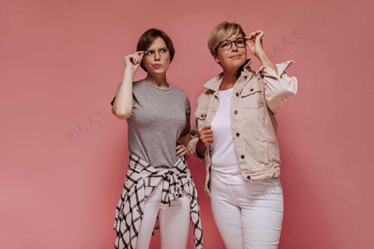 室内短发的两位时尚女性戴着眼镜 穿着白色紧身裤和酷t恤 在孤立的粉色背景上摆姿势老年老年女士