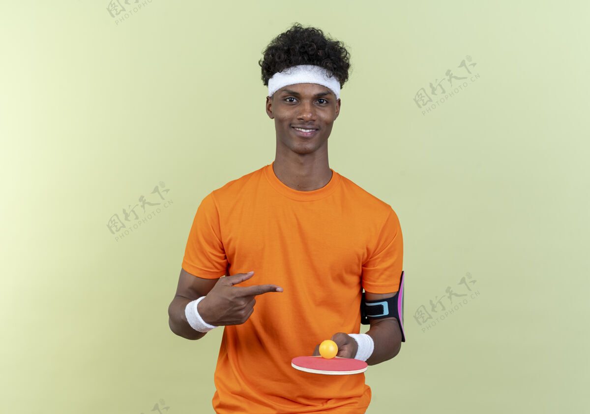 乒乓球高兴的年轻黑人美国运动男子戴着头带和腕带举行和点在乒乓球拍与球隔离在绿色背景球拍穿请