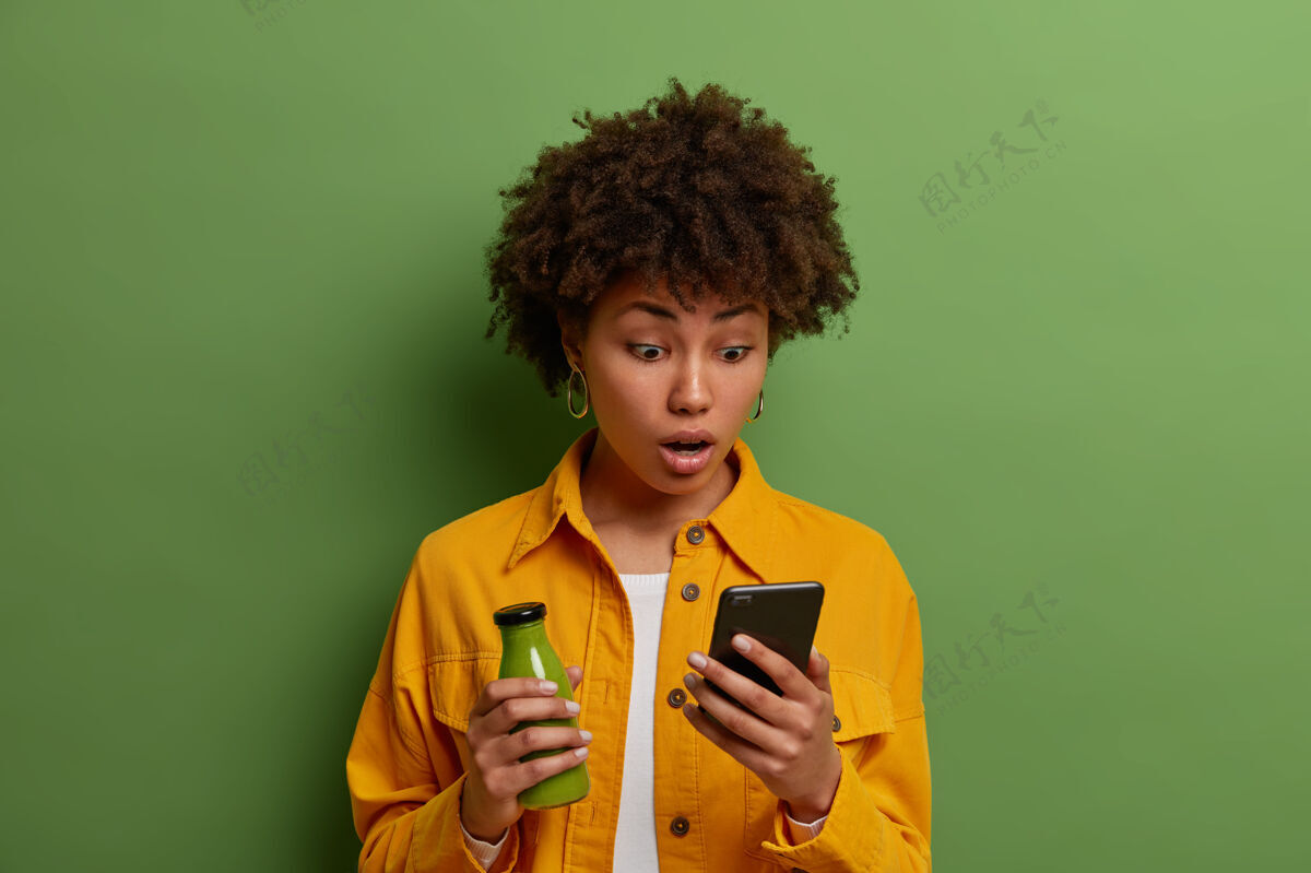 菠菜惊呆了的千禧一代卷发女人盯着智能手机屏幕 读着令人震惊的新闻 喝着菠菜冰沙 使用无线网络 穿着黄色衣服 对着绿色的墙壁摆姿势印象尴尬休息