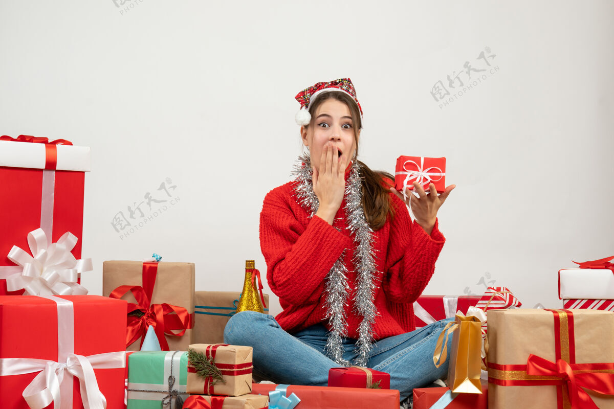 拿着正面图可爱的派对女孩戴着圣诞帽拿着礼物把手放在嘴边围坐着礼物坐着人礼物