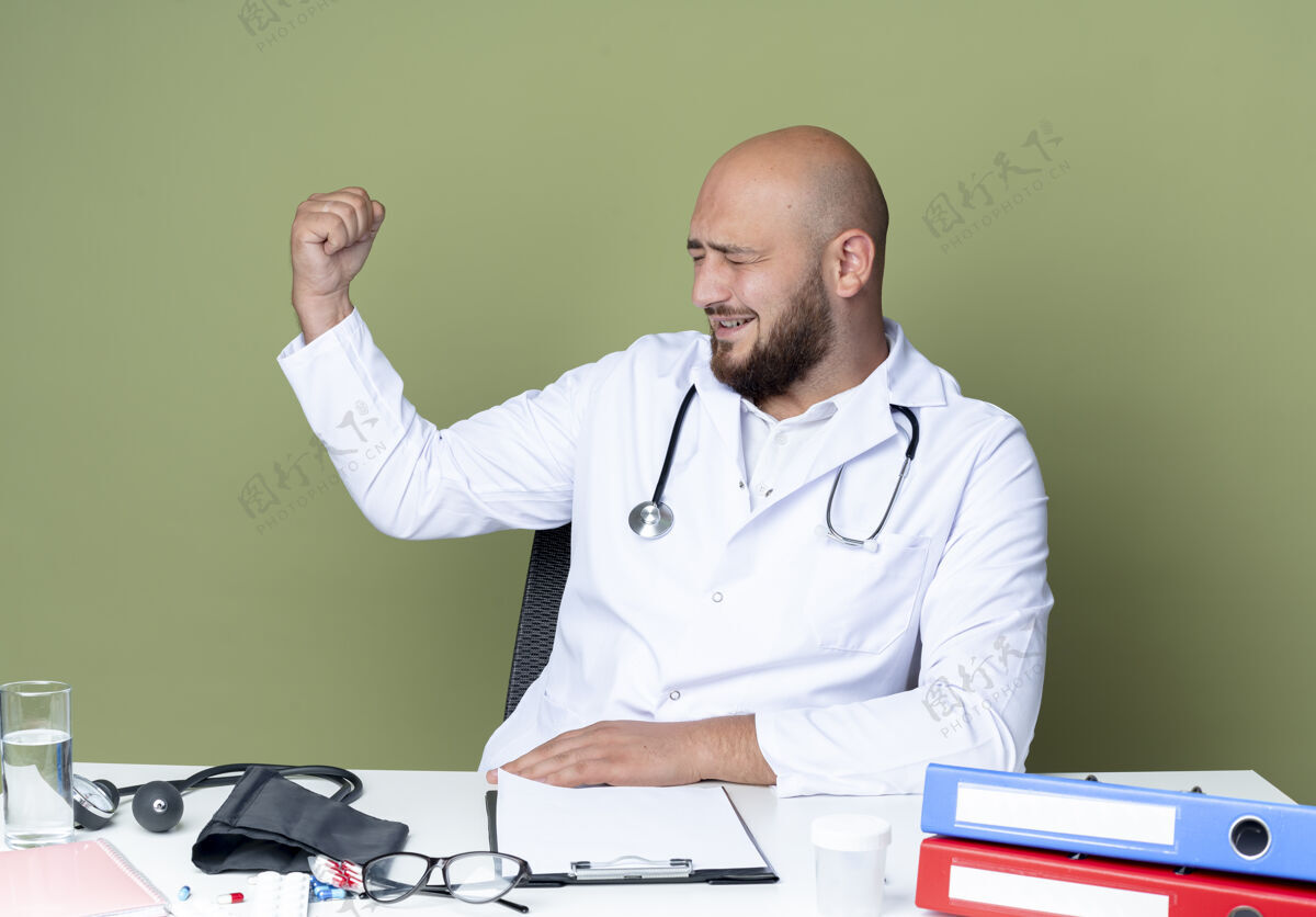 工作高兴的年轻秃头男医生穿着医用长袍和听诊器坐在办公桌前 用医用工具做着强烈的手势隔离在绿色的背景上医疗强壮坐着
