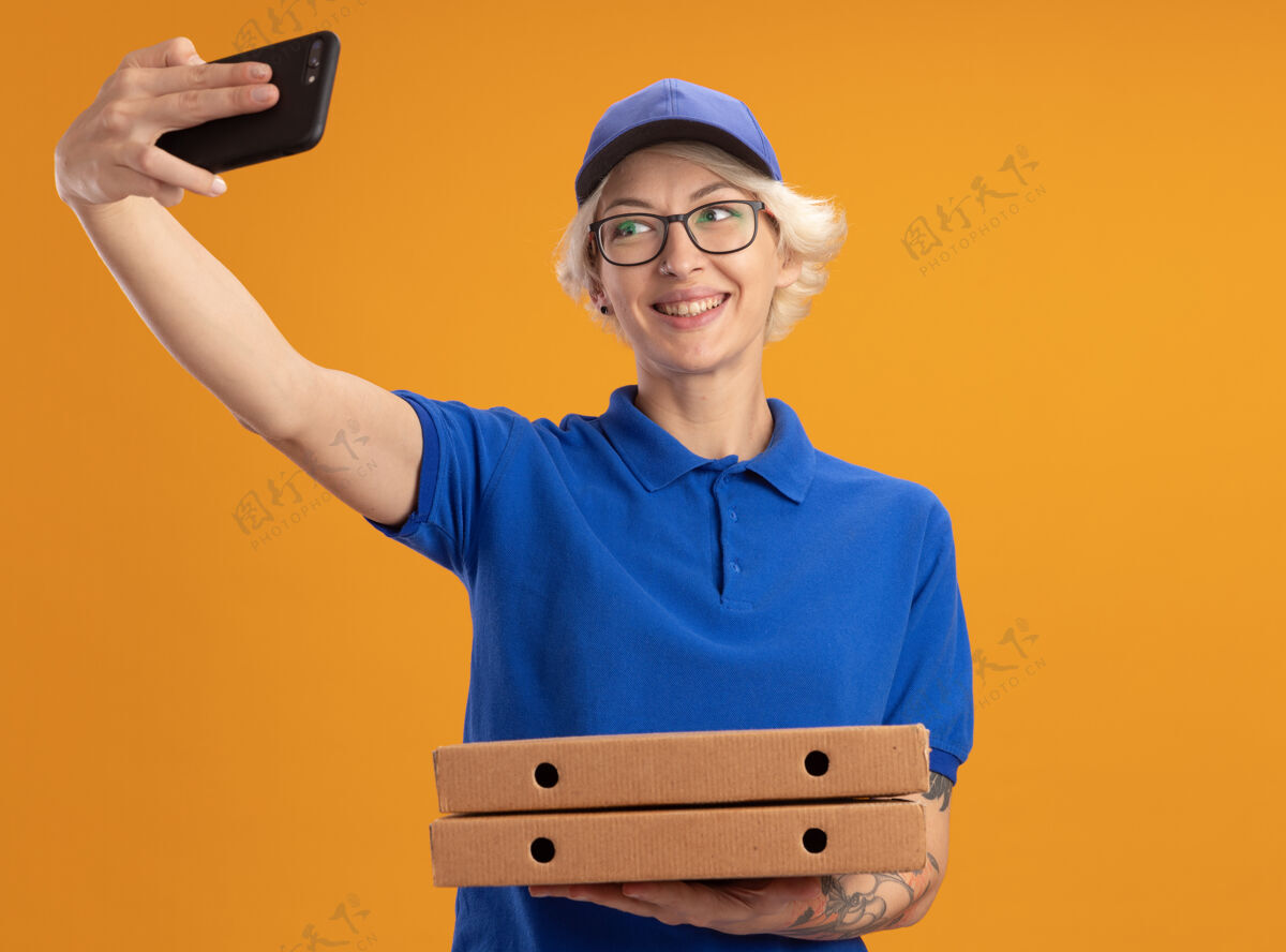送货身穿蓝色制服 戴着眼镜 拿着比萨饼盒 用智能手机在橙色墙上微笑的年轻送货员女士帽子自拍