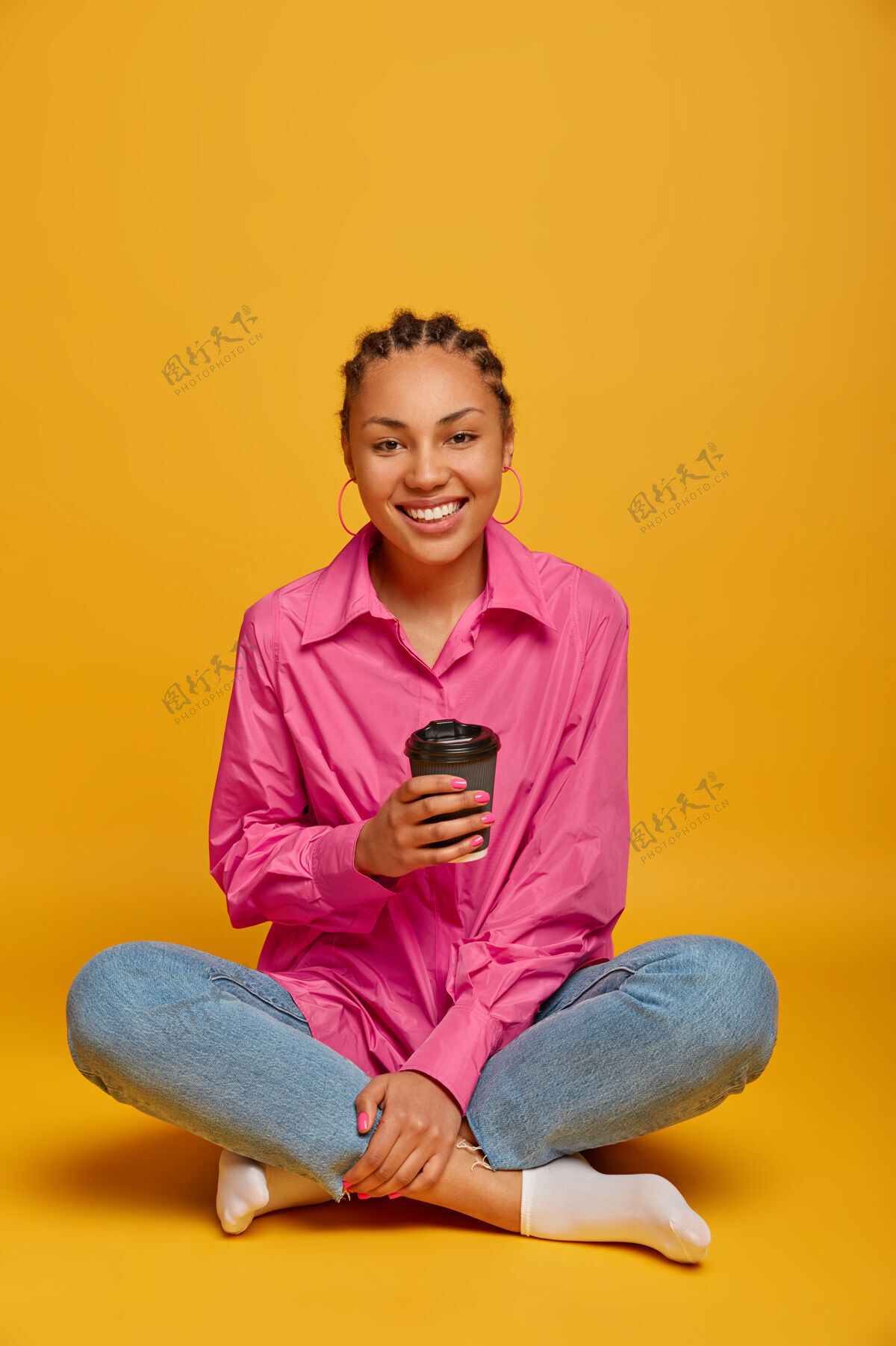 享受快乐的黑皮肤女人坐在莲花姿势的垂直镜头 交叉腿在地板上 喝外卖咖啡 感觉舒服 享受与对话者友好的交谈 隔离在黄色的墙上愉悦摄影满意