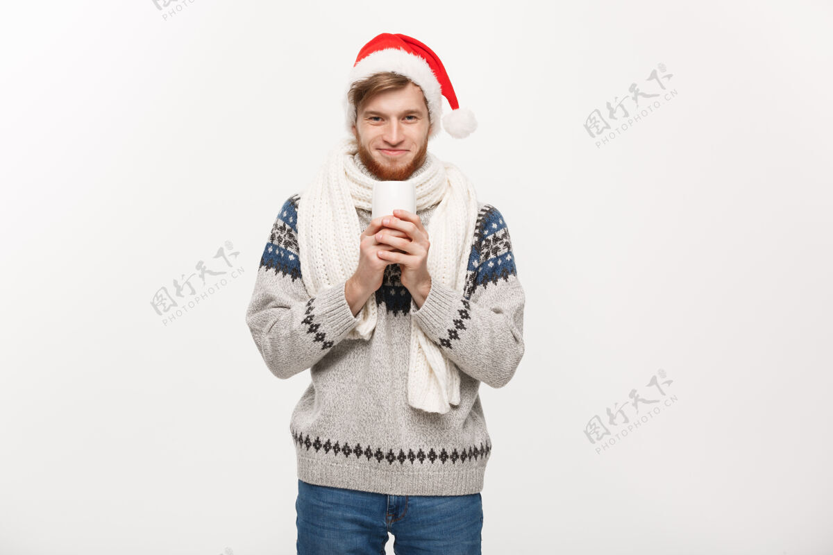 男穿着毛衣 戴着圣诞老人帽 手里拿着一个热咖啡杯的留着胡子的年轻人 孤零零地站在白色的地板上红色咖啡年轻