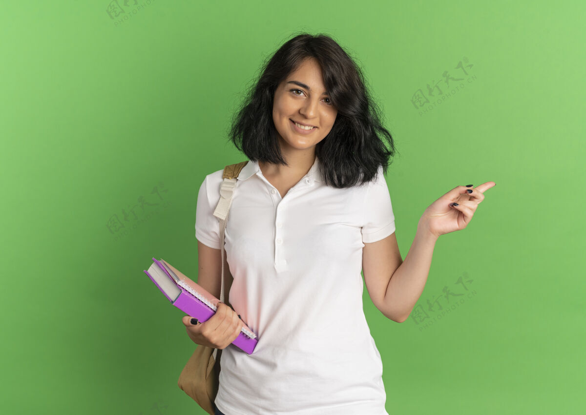 漂亮年轻漂亮的高加索女学生面带微笑 背着书包 站在一旁 手里拿着书 上面有绿色的复印空间微笑包年轻