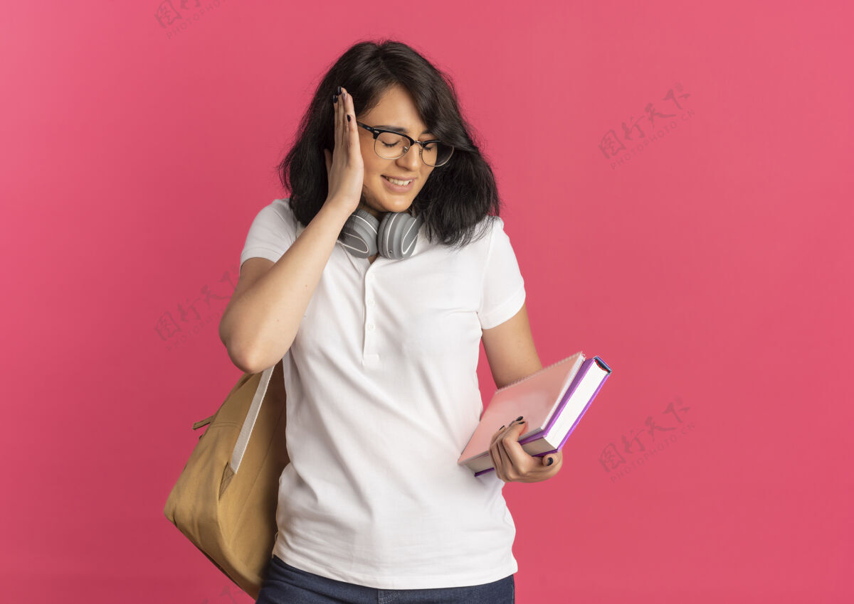 眼镜年轻惹恼漂亮的白人女学生戴着眼镜背上包脖子上戴着耳机用手捂着耳朵拿着书和笔记本粉色带着复印空间穿复制粉红色